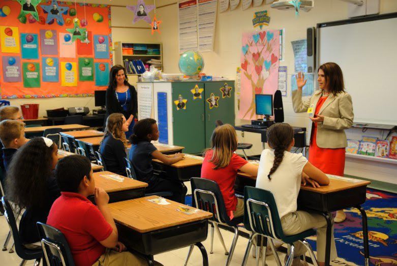 an elementary classroom with six children in desks listening to a presenter at the front of the room