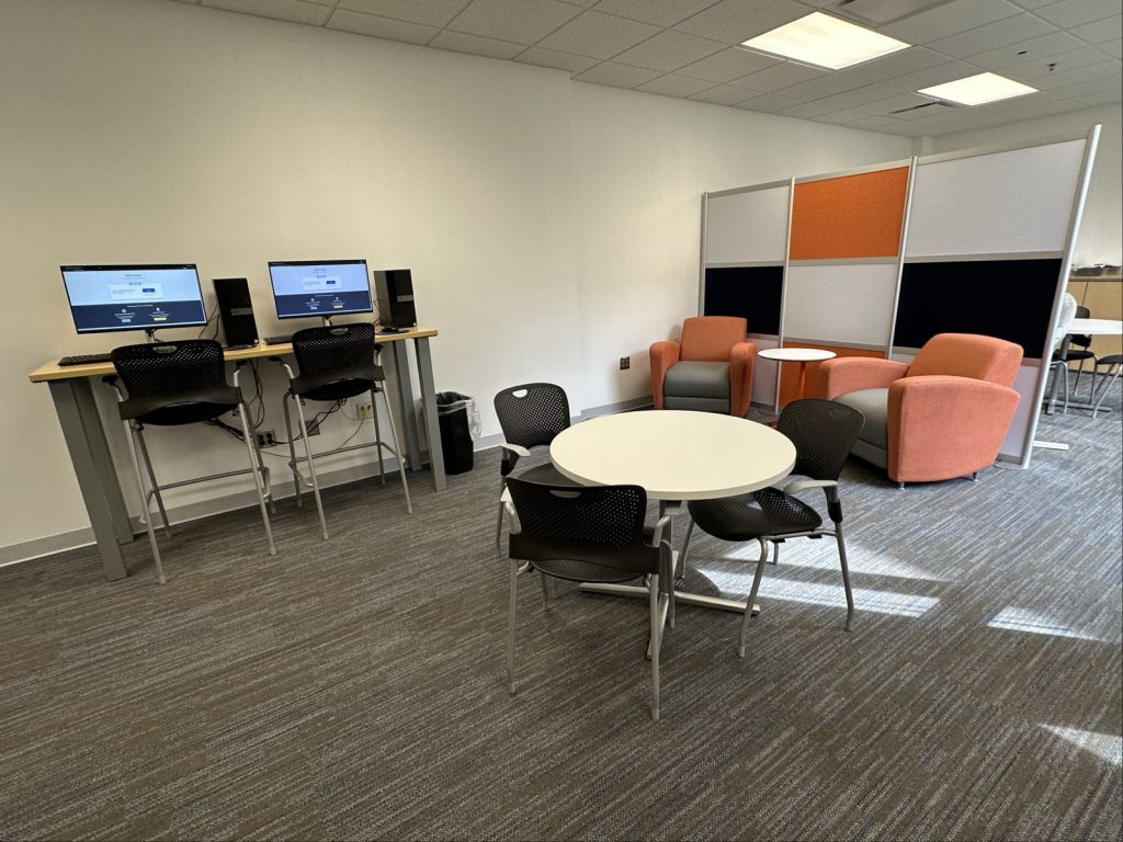 Computers where students can use next to orange couch chairs and a rounded table with chairs.